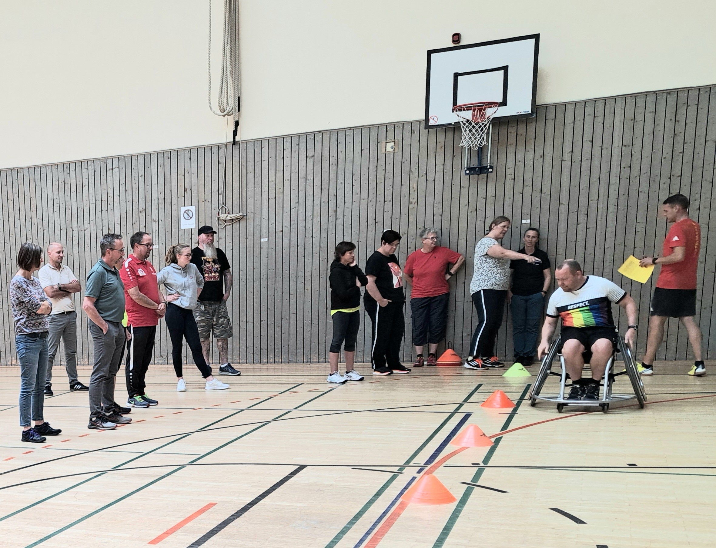 Eine Gruppe von Menschen in einer Sporthalle. Eine Person fährt mit einem Sportrollstuhl durch einen Parcour.